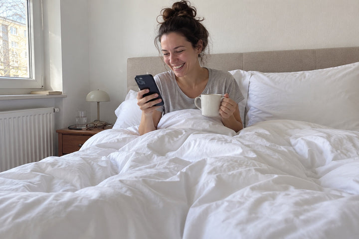 Frau mit Kaffee lacht im Bett mit weißer Baumwoll-Bettwäsche